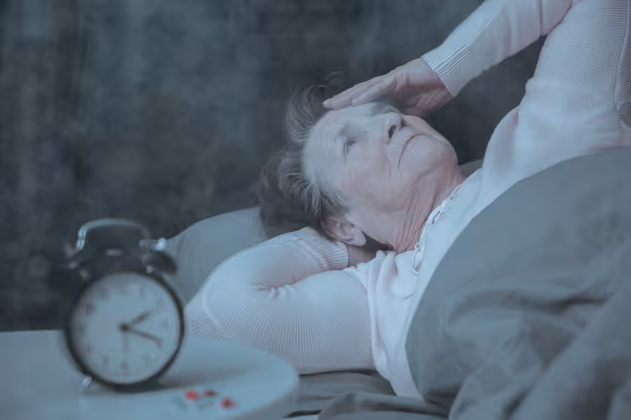 An elderly woman lies awake in bed looking worried, with one hand on her forehead. A clock and some pills are on the bedside table, suggesting insomnia or health concerns.