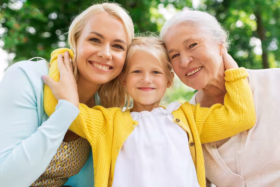 Three generations of women—an older woman, a middle-aged woman, and a young girl—smile and embrace each other outdoors, with green trees blurred in the background.