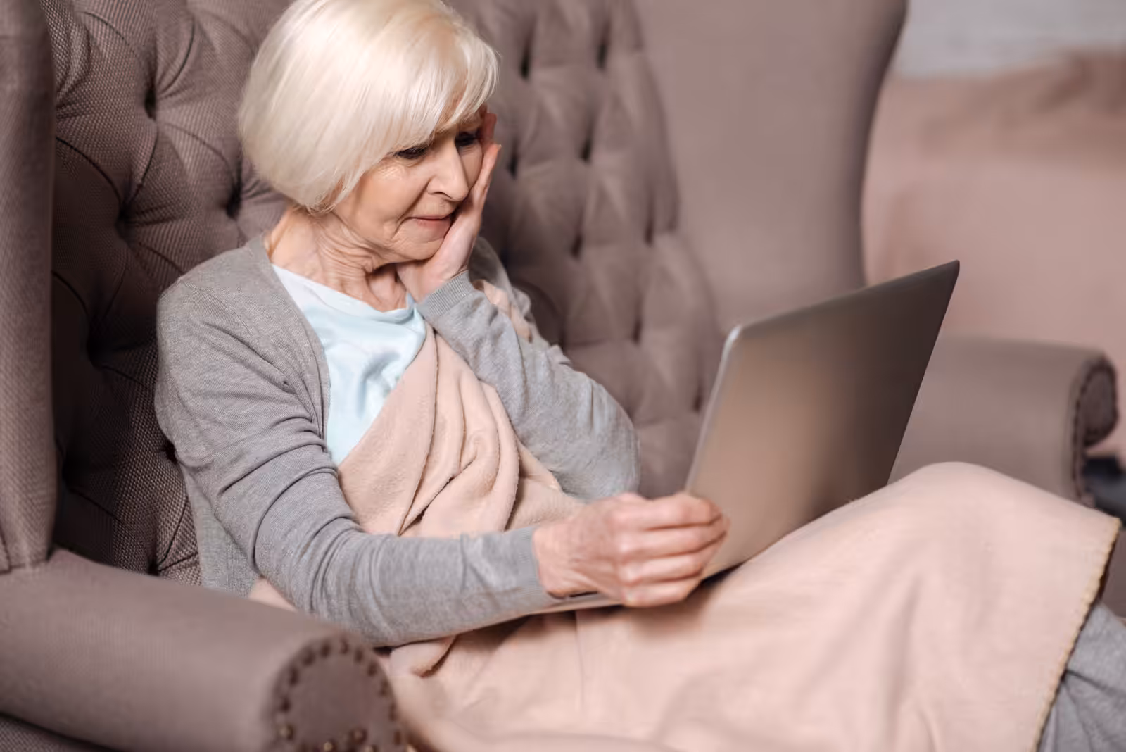 An elderly woman with short white hair sits on a tufted armchair, wrapped in a beige blanket, looking at a laptop on her lap with a thoughtful expression.