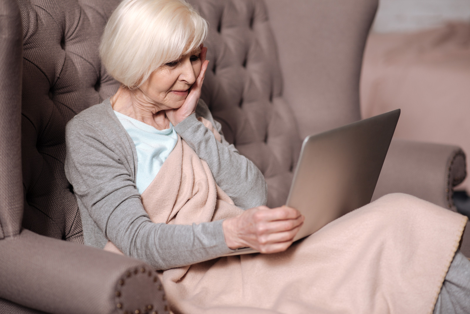 An elderly woman with short white hair sits on a tufted armchair, wrapped in a beige blanket, looking at a laptop on her lap with a thoughtful expression.