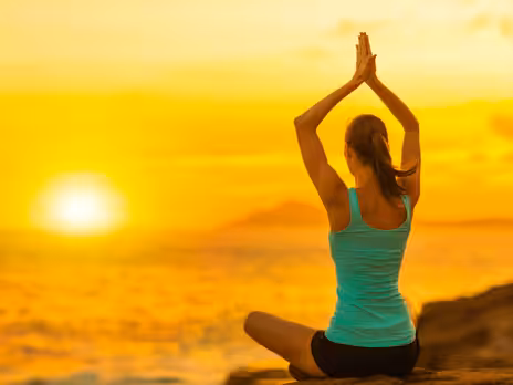 A person sits cross-legged on a rock near the ocean, practicing yoga with hands raised above their head at sunset, with a golden sky and sun in the background.