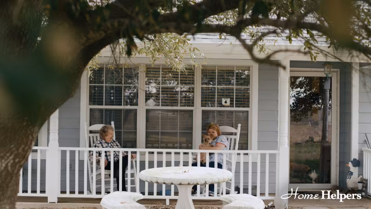 Two women sit on rocking chairs on a front porch, one holding a small dog. A white round table and chairs are in the foreground, and a tree frames the scene. The Home Helpers logo appears in the bottom right corner.
