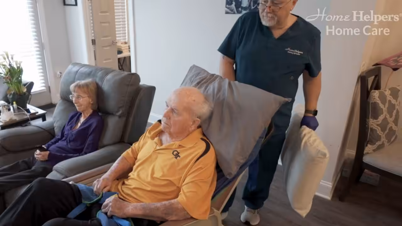 An elderly man sits in a reclining chair with a pillow behind his head, while a caregiver stands beside him holding another pillow. An elderly woman sits nearby in a separate chair. The scene is in a bright living room.