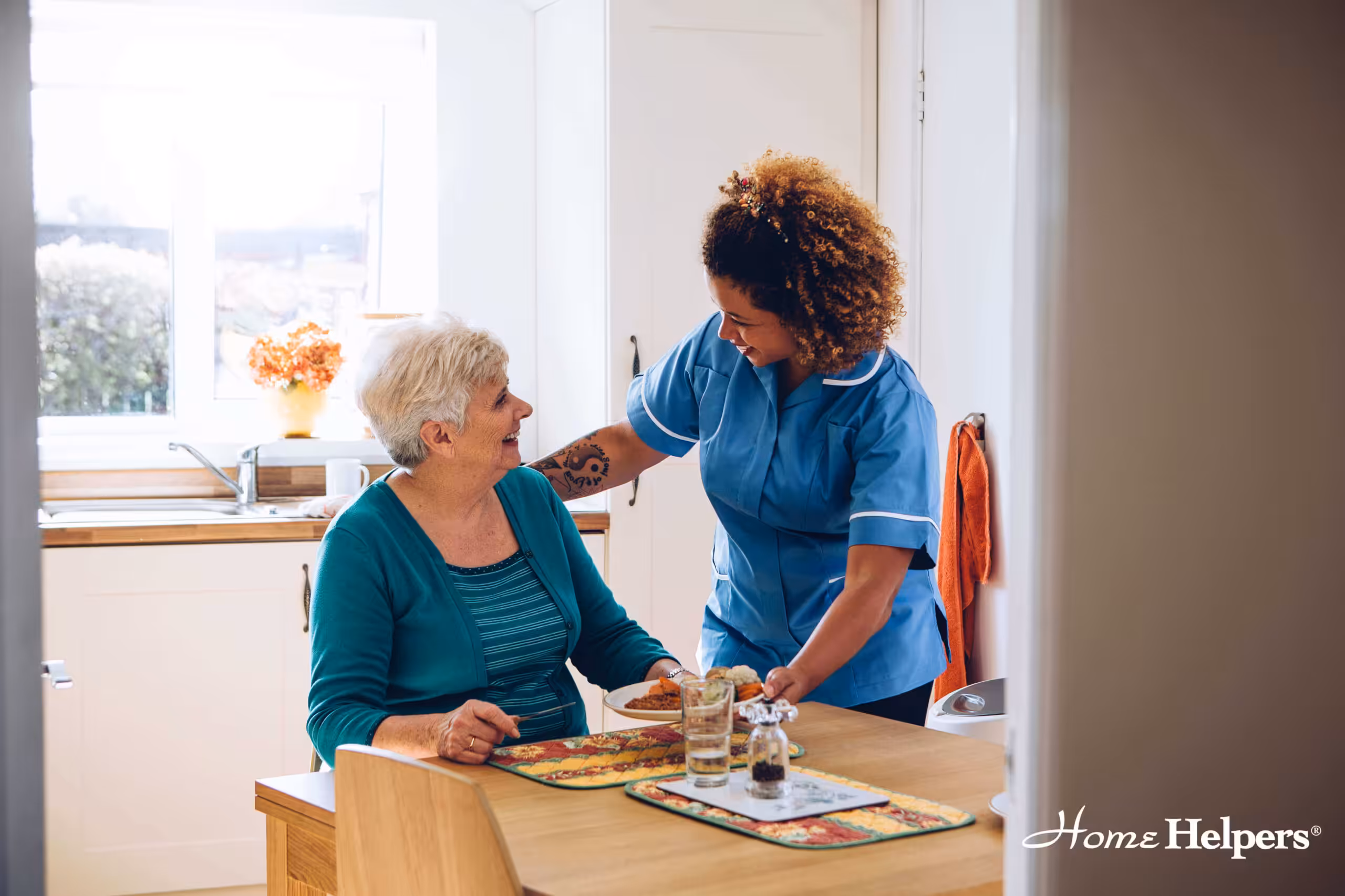 A caregiver in a blue uniform smiles and assists an elderly woman sitting at a kitchen table with a meal. The scene is warm and friendly, with sunlight coming through the window. “Home Helpers” is written in the bottom right corner.