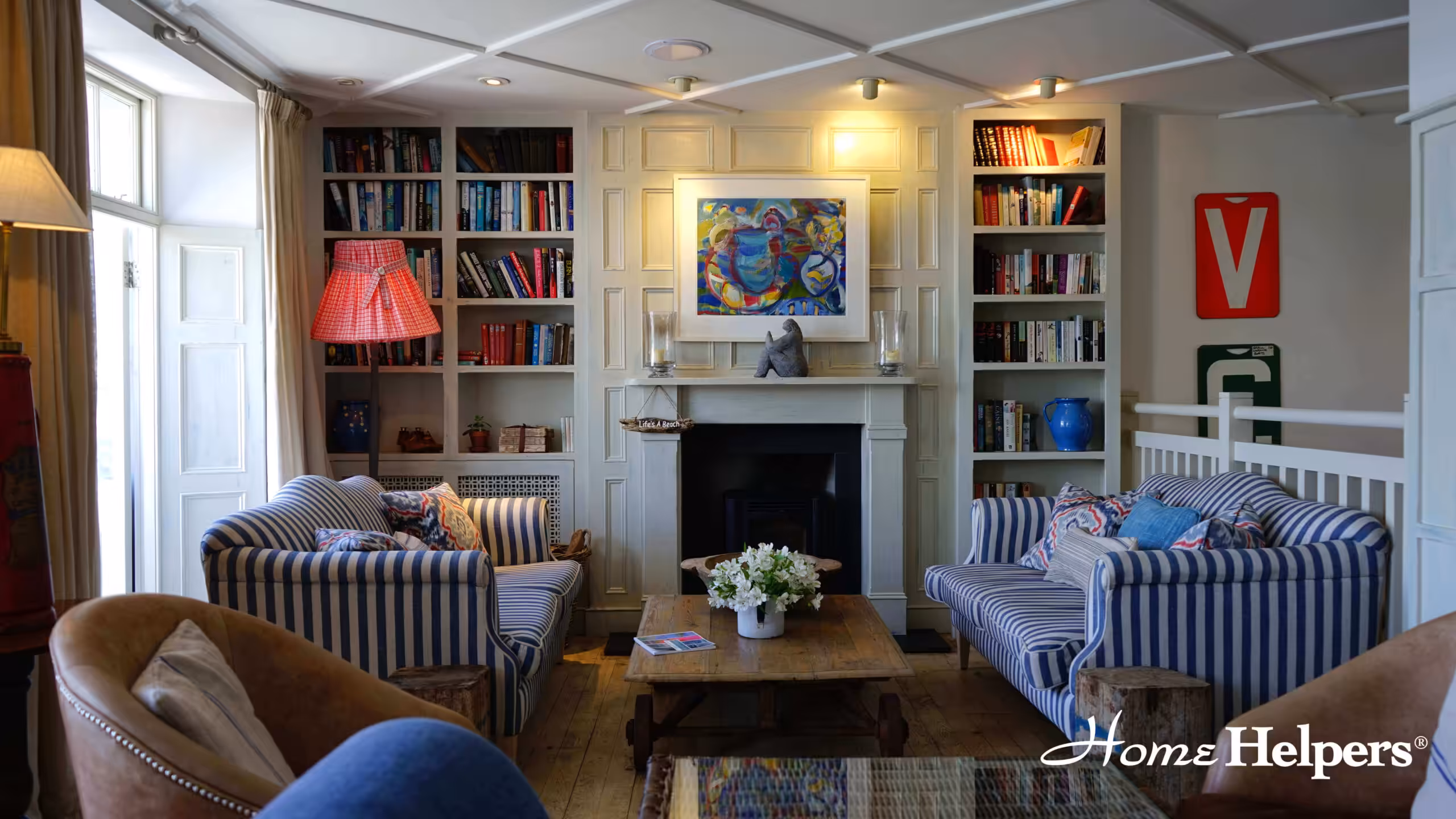 A cozy living room with striped sofas, a wooden coffee table, bookshelves filled with books, and art above a fireplace. Warm light fills the room, and flowers decorate the table. The Home Helpers logo is in the corner.