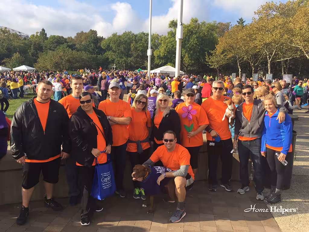 A group of people in matching orange shirts pose together outdoors at a crowded event. They are smiling, some wearing sunglasses, with trees and a large crowd in the background. A dog in a purple shirt sits in front of them.