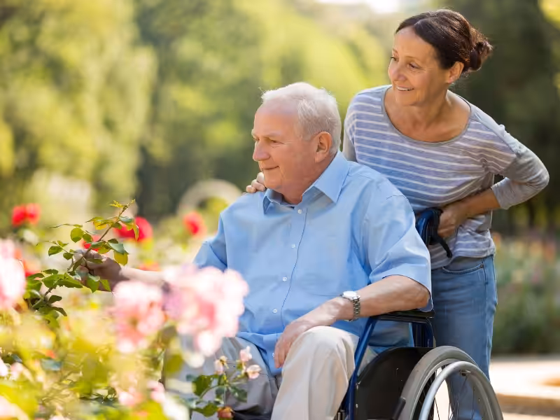An elderly man in a wheelchair enjoys a garden with blooming flowers, accompanied by a smiling woman standing behind him. The scene is bright and outdoors, conveying a sense of care and companionship.