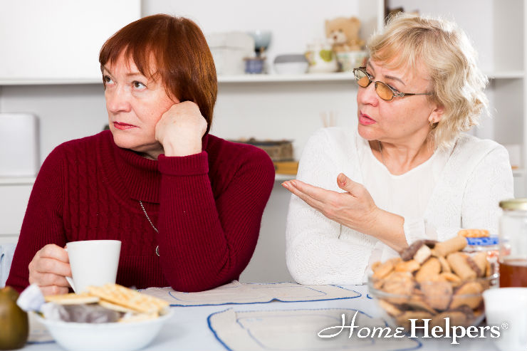 Two older women sit at a kitchen table with snacks and drinks. One looks upset and turns away, while the other gestures toward her as if talking or explaining. The Home Helpers logo is visible in the corner.