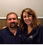 A man and woman sit closely together, smiling at the camera. They both wear dark blue shirts with a small embroidered logo, and are seated in front of a beige and white wall.