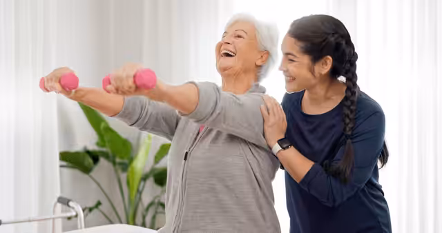 An older woman smiles as she lifts pink dumbbells while a younger woman beside her offers support and encouragement, both appearing happy and energetic in a bright room with plants.