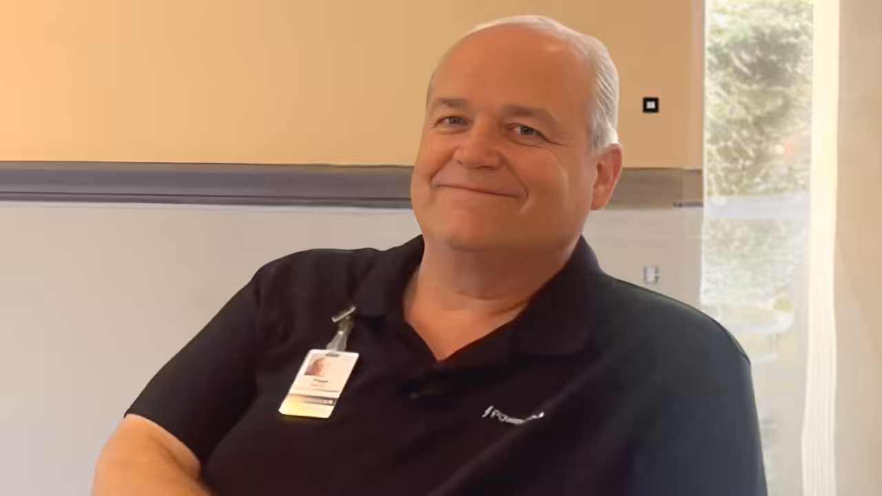 A smiling older man with short gray hair sits indoors, wearing a black collared shirt and an ID badge clipped to his chest. A window with light curtains and greenery outside is visible in the background.