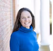 A woman with long brown hair, wearing a bright blue turtleneck sweater, stands smiling next to a brick wall with white columns in the background.