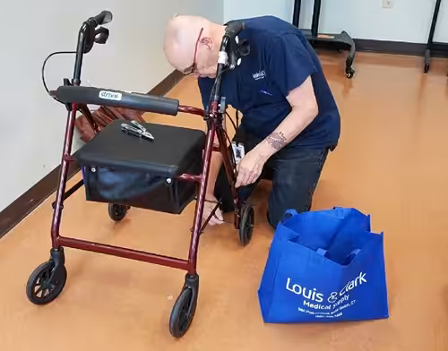 A man wearing glasses and a blue shirt kneels to repair or inspect the wheel of a red walker. A blue Louis & Clark Medical Supply tote bag sits on the floor nearby.