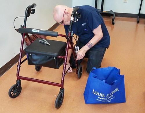 A man wearing glasses and a blue shirt kneels to repair or inspect the wheel of a red walker. A blue Louis & Clark Medical Supply tote bag sits on the floor nearby.
