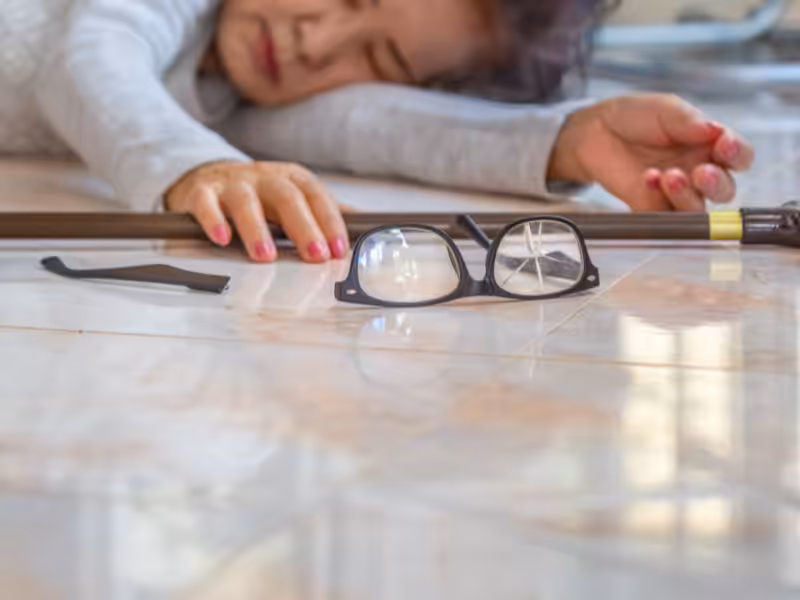 An elderly person lies on the floor reaching for a walking cane. Eyeglasses are on the floor nearby, out of reach. The scene suggests the person has fallen.