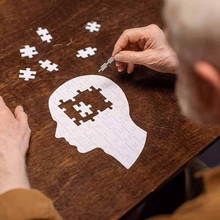 An older person assembles a puzzle shaped like a human head on a table, with one piece being placed in the brain area, symbolizing mental health or memory.
