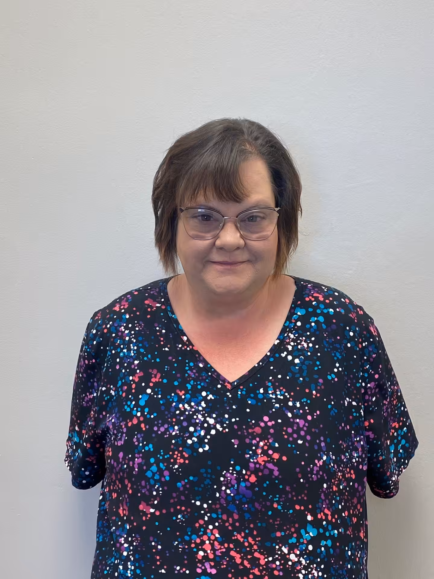 A woman with short brown hair and glasses stands against a plain light-colored wall, wearing a black shirt with a colorful splatter pattern. She is looking at the camera with a slight smile.