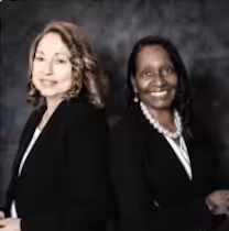 Two women wearing black blazers and white tops stand side by side, smiling at the camera against a dark, textured background.