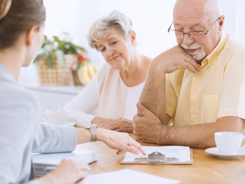 An older couple sits at a table, looking thoughtfully at a clipboard as a woman in a blazer explains documents to them. A coffee cup and papers are on the table.