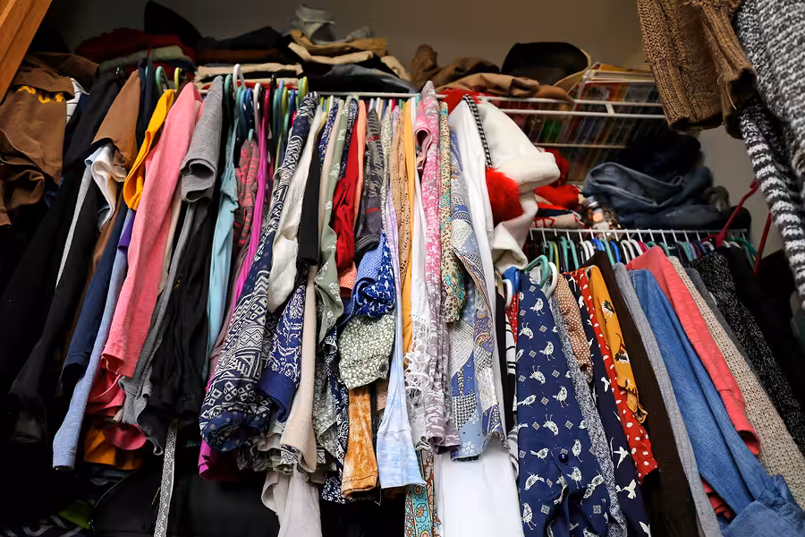 A crowded closet filled with various colorful clothes hanging on hangers, with some garments draping over each other and shelves above holding bags and folded items.