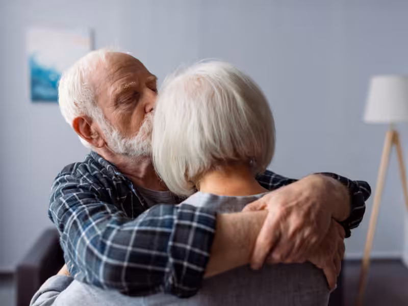 An elderly man with white hair and a beard hugs an elderly woman with short white hair, gently kissing her head. They are indoors, sharing an affectionate and comforting moment.