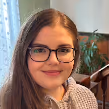 A young woman with long brown hair and glasses is smiling. She is wearing a light-colored knitted sweater and is indoors, with a plant and wooden furniture in the background. Light is coming through a window with sheer curtains.