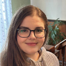 A young woman with long brown hair and glasses is smiling. She is wearing a light-colored knitted sweater and is indoors, with a plant and wooden furniture in the background. Light is coming through a window with sheer curtains.