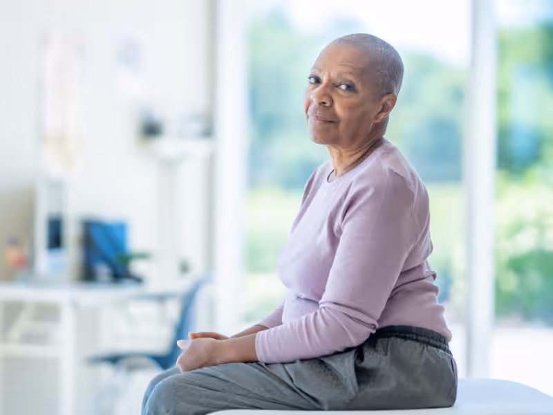 An older woman with short hair and a light purple sweater sits on a medical exam table, looking at the camera with a gentle expression. The background is bright and appears to be a doctors office.