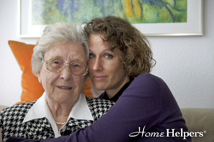 An elderly woman with glasses and a younger woman with curly hair sit close together on a couch, smiling gently. The younger woman hugs the older woman. The words “Home Helpers” appear in the bottom right corner.