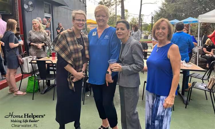 Four women stand and smile at an outdoor gathering with tables, tents, and people in the background. The “Home Helpers” logo and “Clearwater, FL” text appear in the bottom left corner.