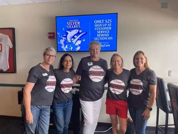 Five women stand together smiling, wearing matching Community Warrior T-shirts in a room. Behind them is a blue sign advertising a Silver Sharks event for $25 at customer service behind Section 105.