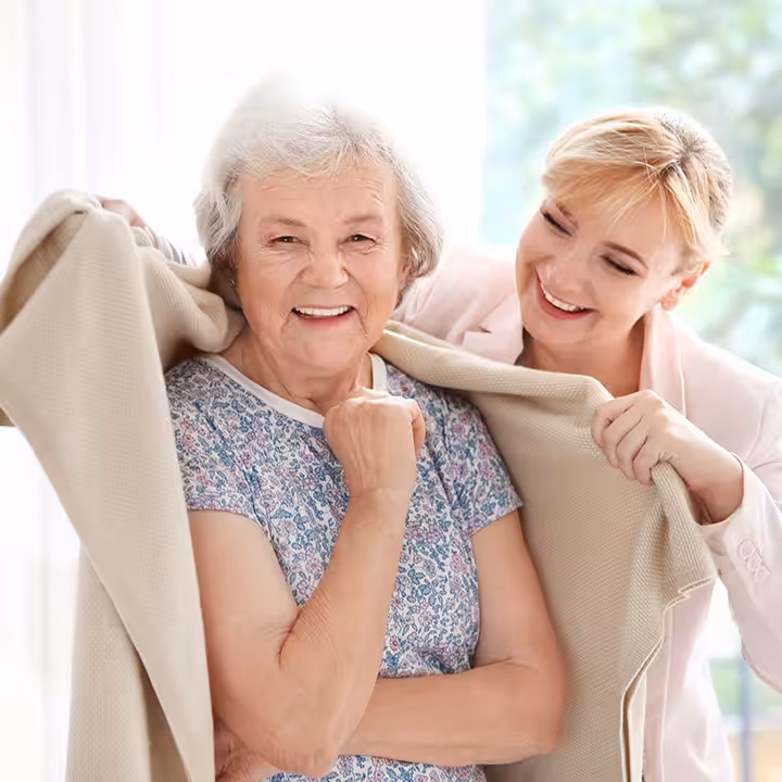 A smiling older woman with gray hair is wrapped in a beige blanket by a younger woman beside her. Both women are smiling and appear happy in a bright, sunlit room with a green, blurred background.