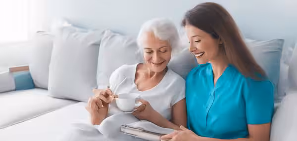 An elderly woman sits on a couch, smiling and holding a cup, while a younger woman in blue scrubs sits beside her, showing her something on a clipboard. Both appear happy and engaged.