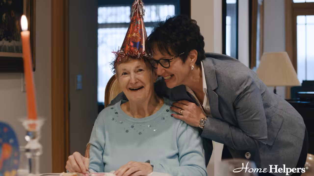 An elderly woman in a party hat smiles while seated at a table, as a woman in glasses and a blazer leans in close, both appearing happy. A lit candle is visible in the foreground. Home Helpers logo is in the corner.