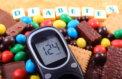 A glucose meter displaying 124 mg/dL is surrounded by assorted candies, chocolates, and cookies. In the background, letter blocks spell out DIABETES.