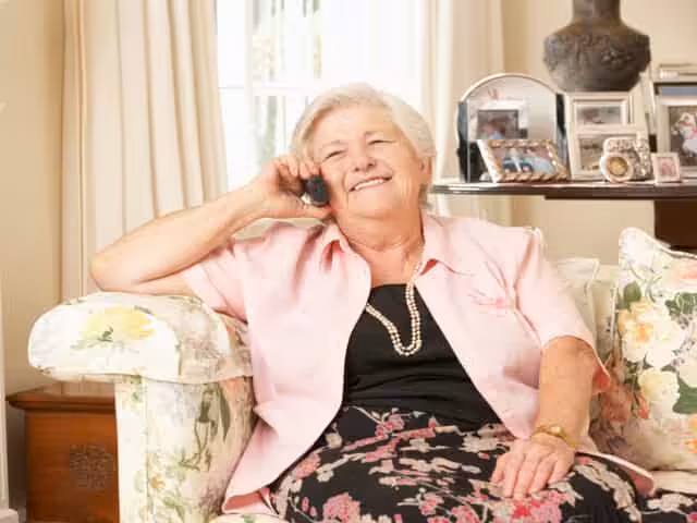 An elderly woman with grey hair, wearing a pink blouse and patterned skirt, sits on a floral armchair, smiling while talking on a phone. Family photos and decor are visible on a table behind her.
