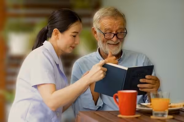 A smiling older man with glasses reads a book with a young woman beside him, both looking at the pages. They sit at a table with a red mug, orange juice, and a plate of food.