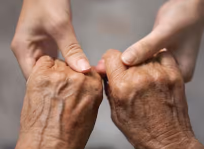 Two younger hands gently hold the thumbs of two older, wrinkled hands, conveying support, care, and connection across generations.