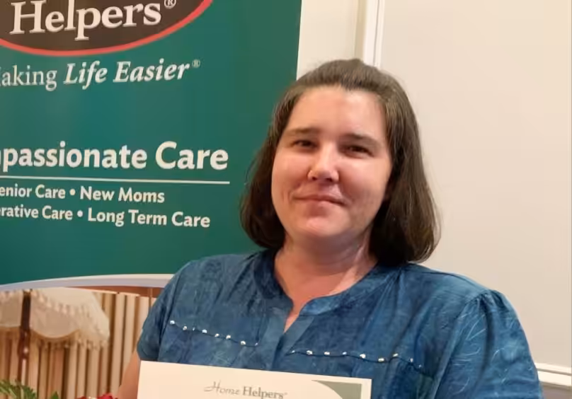 A woman with shoulder-length brown hair holds a certificate and stands in front of a green Home Helpers banner that mentions compassionate care for senior care, new moms, recuperative care, and long-term care.