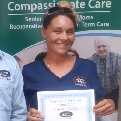 A woman in blue scrubs smiles while holding an Employee of the Month certificate for August 2017. The certificate has the Home Helpers logo. A banner behind her reads Compassionate Care and features images of seniors.