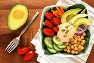 A bowl filled with sliced avocado, cherry tomatoes, cucumber, spinach, carrots, red onion, chickpeas, and a scoop of hummus. Half an avocado, a fork, and grape tomatoes are on a wooden surface nearby.