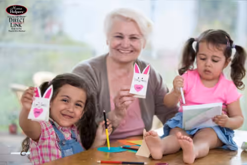 An older woman and two young girls sit at a table doing crafts. One girl and the woman hold up paper bunny crafts, while the other girl colors in a book. All are smiling and enjoying the activity together.