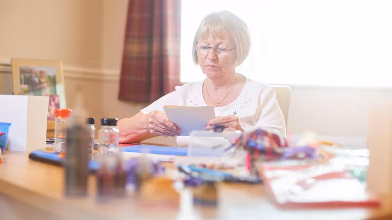 An elderly woman sits at a table with craft supplies, holding paper and scissors, focused on her work. The room is softly lit, with various art materials and a framed photo visible in the background.