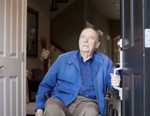 An elderly man in a blue jacket sits in a wheelchair by an open door, looking thoughtfully outside. The scene appears to be in the entrance of a home.