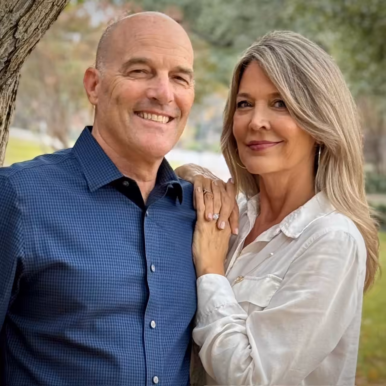A man in a blue shirt and a woman in a white blouse stand close together outdoors, smiling at the camera. The woman has her hand on the mans chest, and trees and greenery are visible in the background.