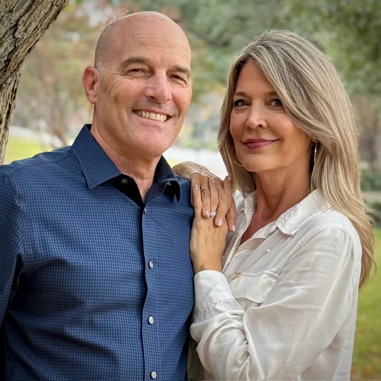 A man in a blue shirt and a woman in a white blouse stand close together outdoors, smiling at the camera. The woman has her hand on the mans chest, and trees and greenery are visible in the background.