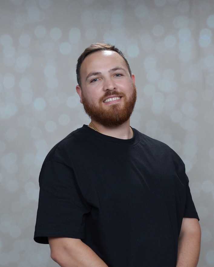A man with a short beard and short hair smiles at the camera. He is wearing a plain black t-shirt and stands in front of a light, blurry background with circular bokeh patterns.