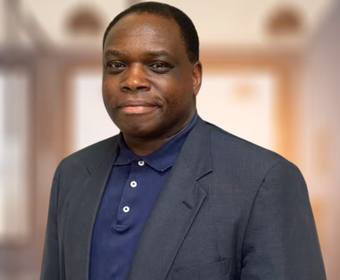 A man wearing a dark blazer over a navy polo shirt stands indoors in front of a softly blurred background with warm lighting. He has short hair and a slight smile.