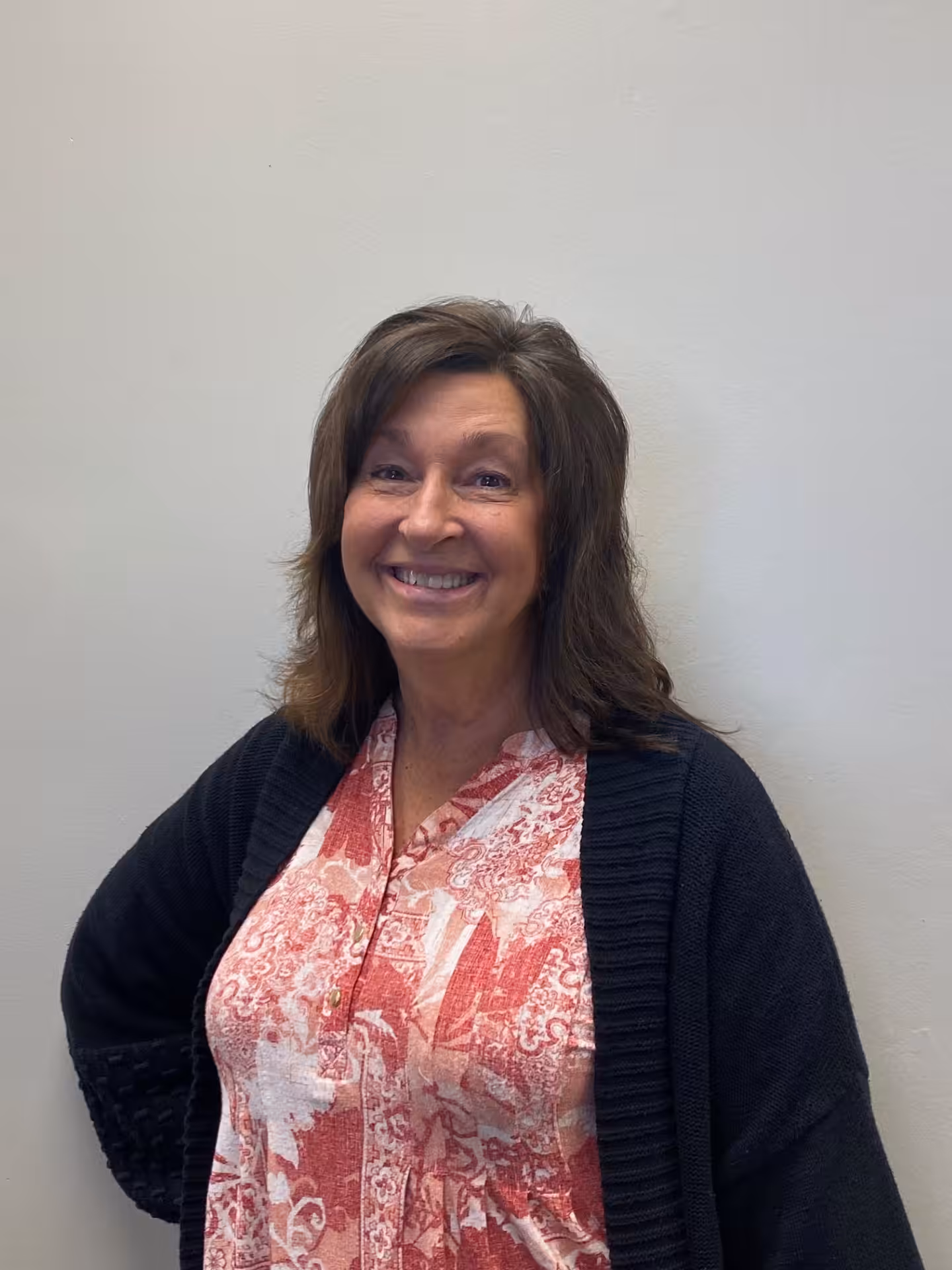 A woman with shoulder-length brown hair smiles at the camera. She is wearing a pink and white patterned blouse and a black cardigan, standing in front of a plain light-colored wall.