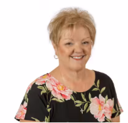 A smiling older woman with short, light hair wearing a black top with large pink floral patterns and a cross necklace, photographed against a plain white background.